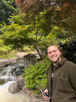 Luke Townrow is smiling with a phone in his hand. He stands in front of a small waterfall surrounded by rocks, trees and a heron.