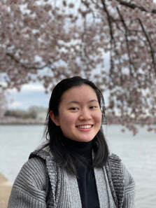 Melodie Qian is smiling and wearing gray winter clothes. The blurred background shows a tree branch and a body of water.